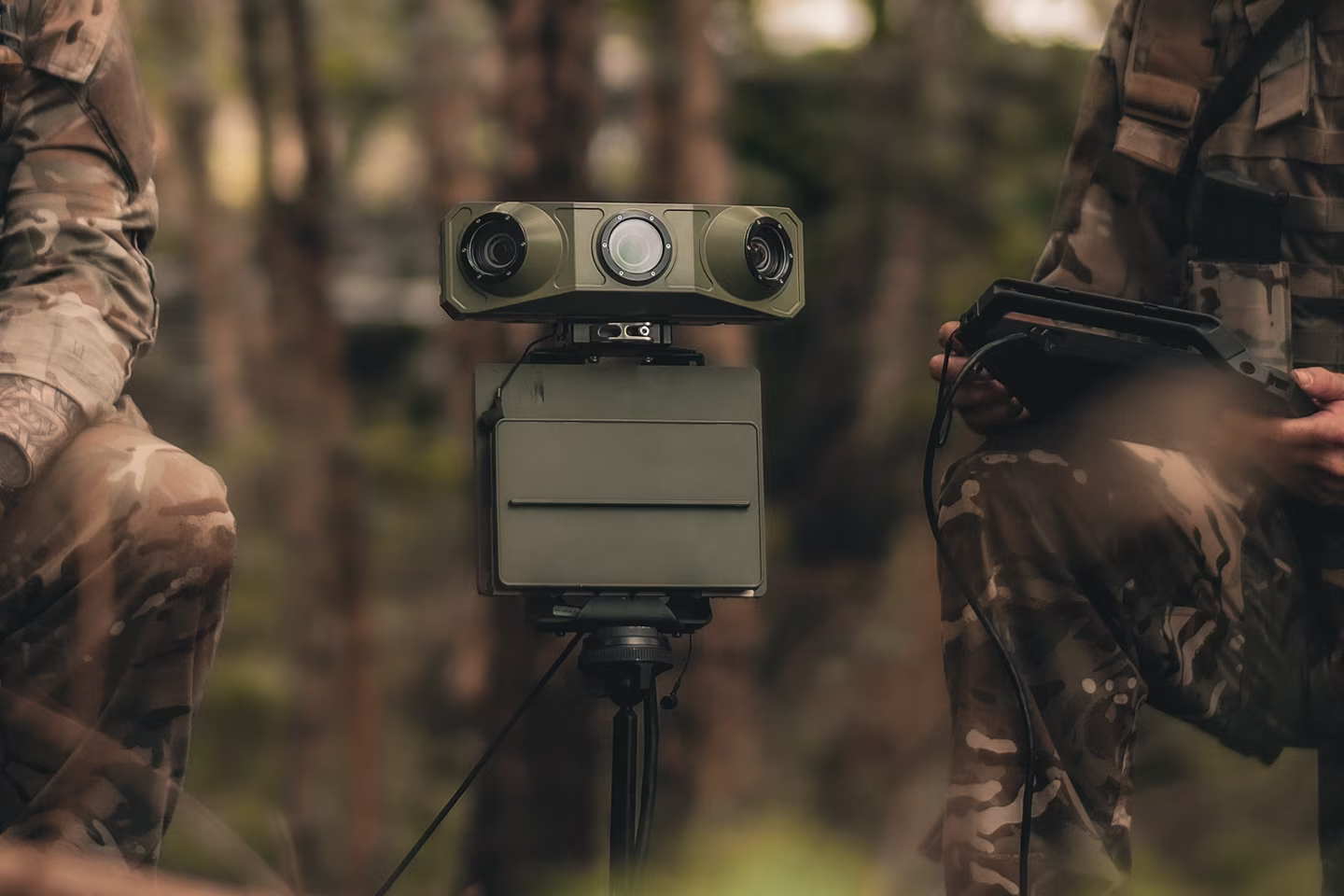 Military device with cameras on tripod between two soldiers in camouflage holding equipment.