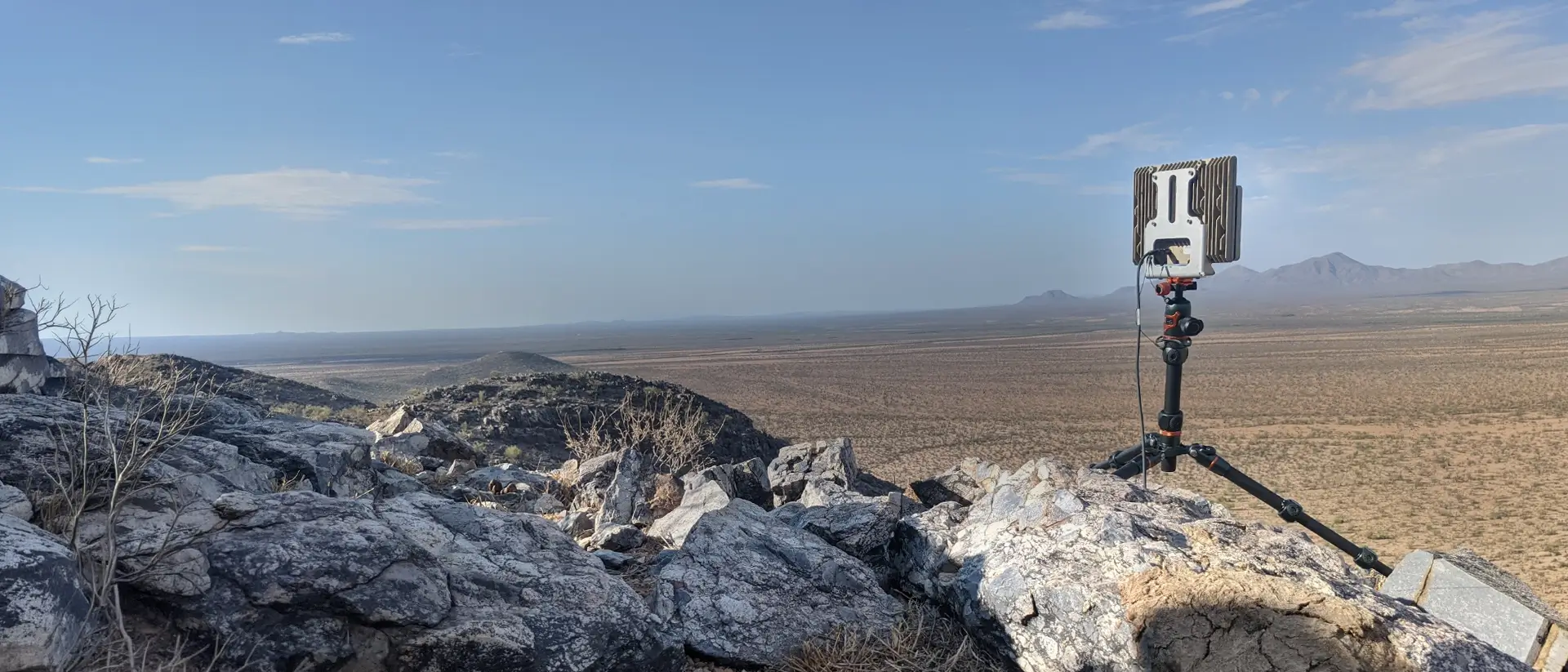 Tripod-mounted device on rocky hill overlooking an expansive desert landscape with distant mountains.