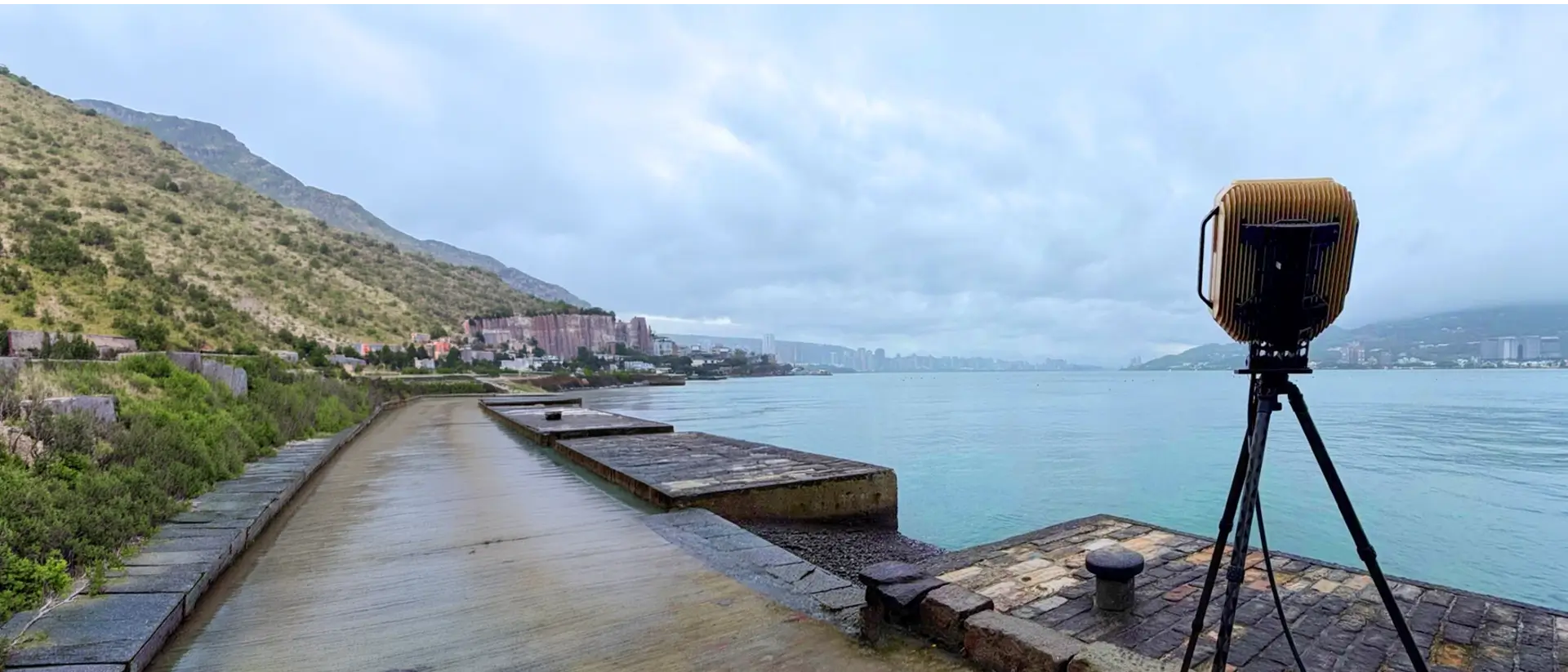 Waterfront path beside calm bay with mountains, buildings, and a device on a tripod in the foreground.