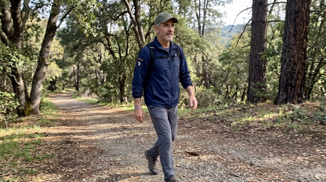 Man wearing a cap and jacket hiking on a forest trail surrounded by trees and greenery.