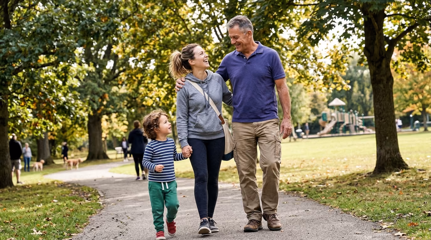 Family of three walking and smiling on a park path surrounded by trees and grass.