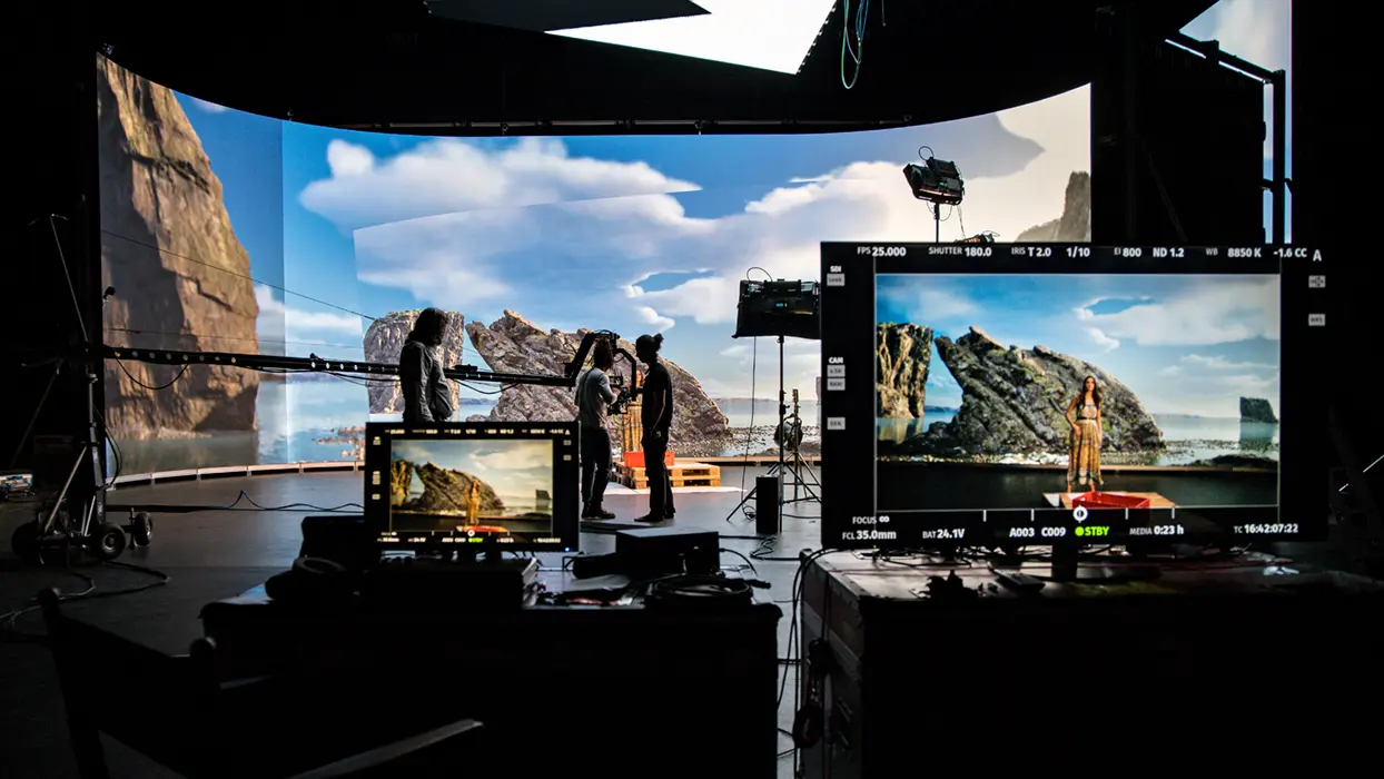 Film crew in front of an LED backdrop showing a coastal landscape, with control monitors in the HALOSTAGE studio.
