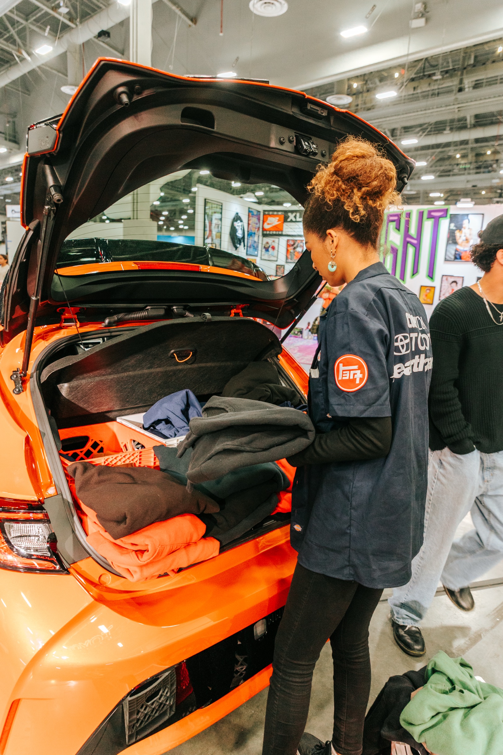 Visitor browsing merch staged in the open trunk of an orange Toyota