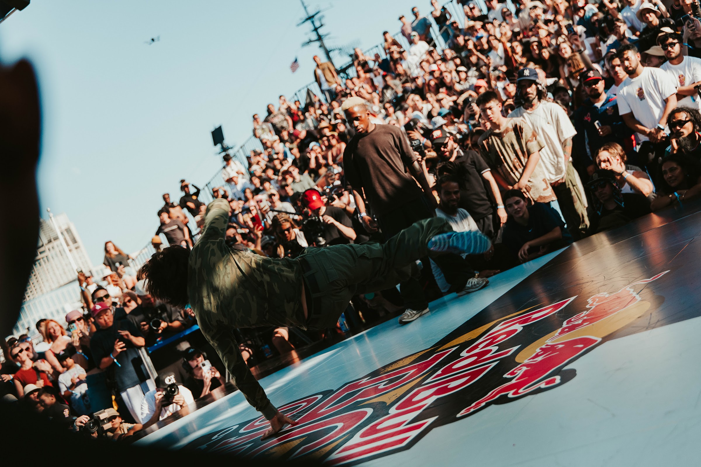 B-boy mid-freeze on the Red Bull BC One floor in front of packed bleachers on San Diego's Broadway Pier