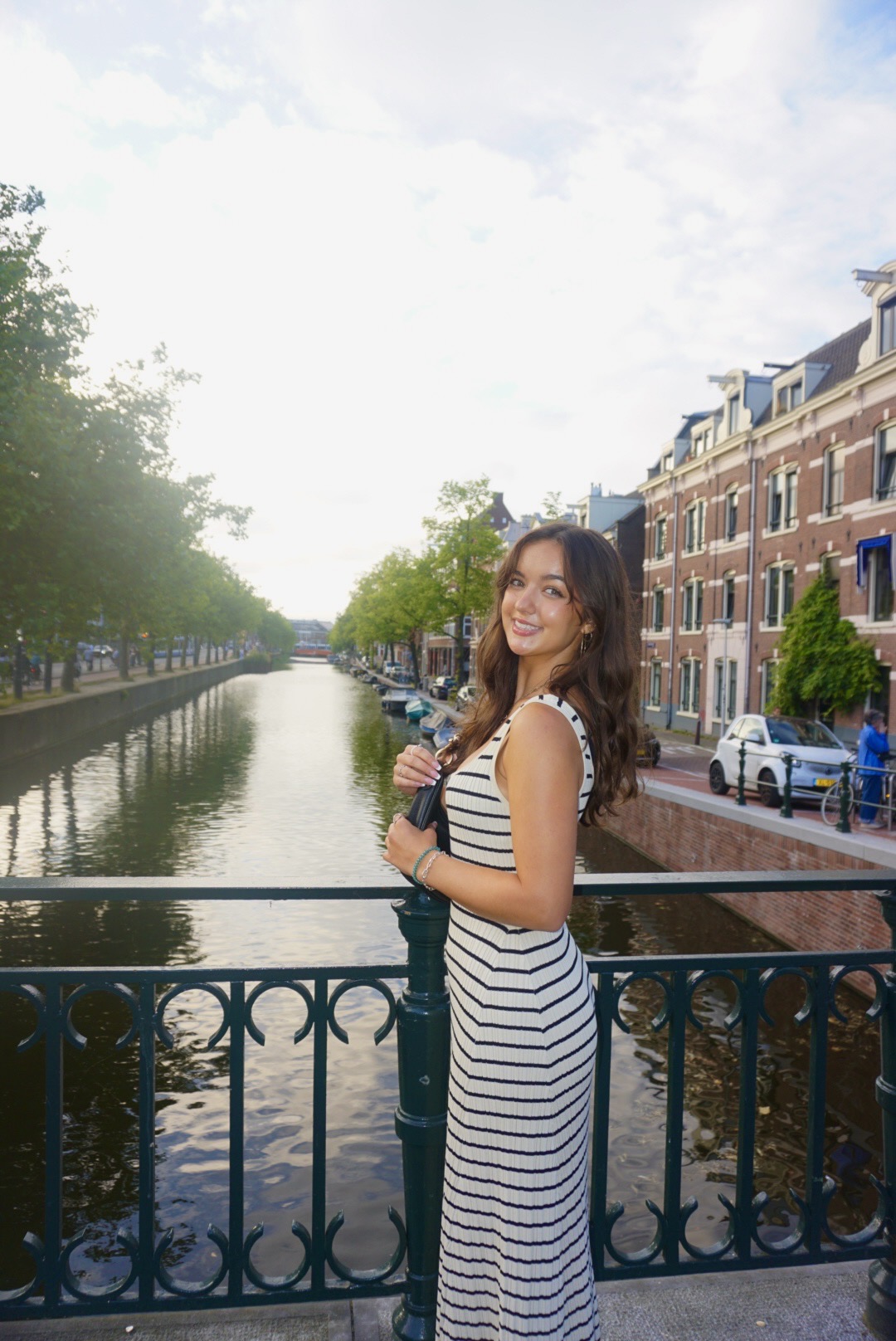 A young woman with long brown hair in a sleeveless white dress with black horizontal stripes stands smiling by a green wrought iron railing over a canal lined with trees and buildings.