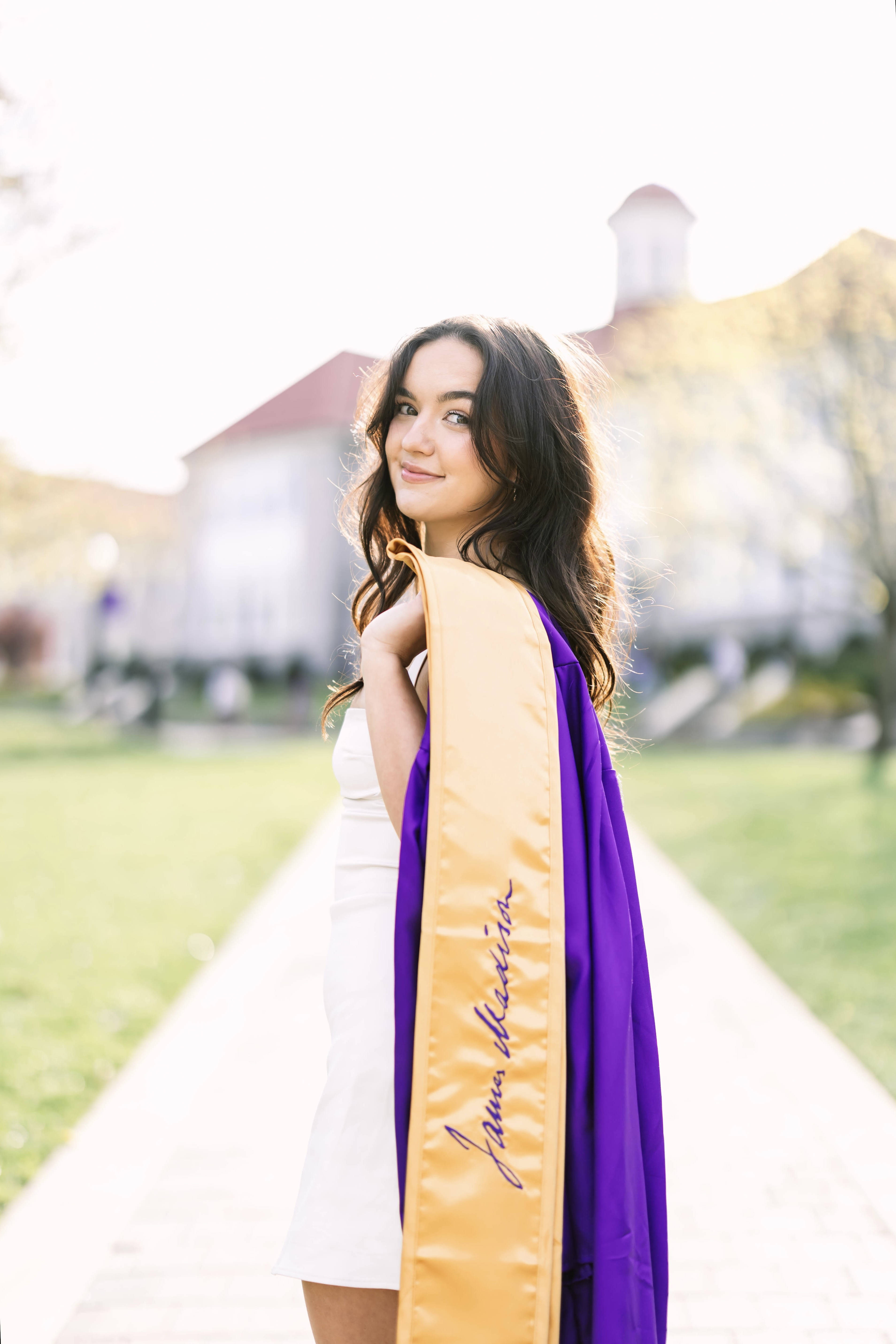 Young woman with long dark hair standing outdoors on a paved path, wearing a white dress and holding a purple and gold graduation stole with the name 'Janna Martin' embroidered on it, smiling softly with a blurred building and greenery in the background.