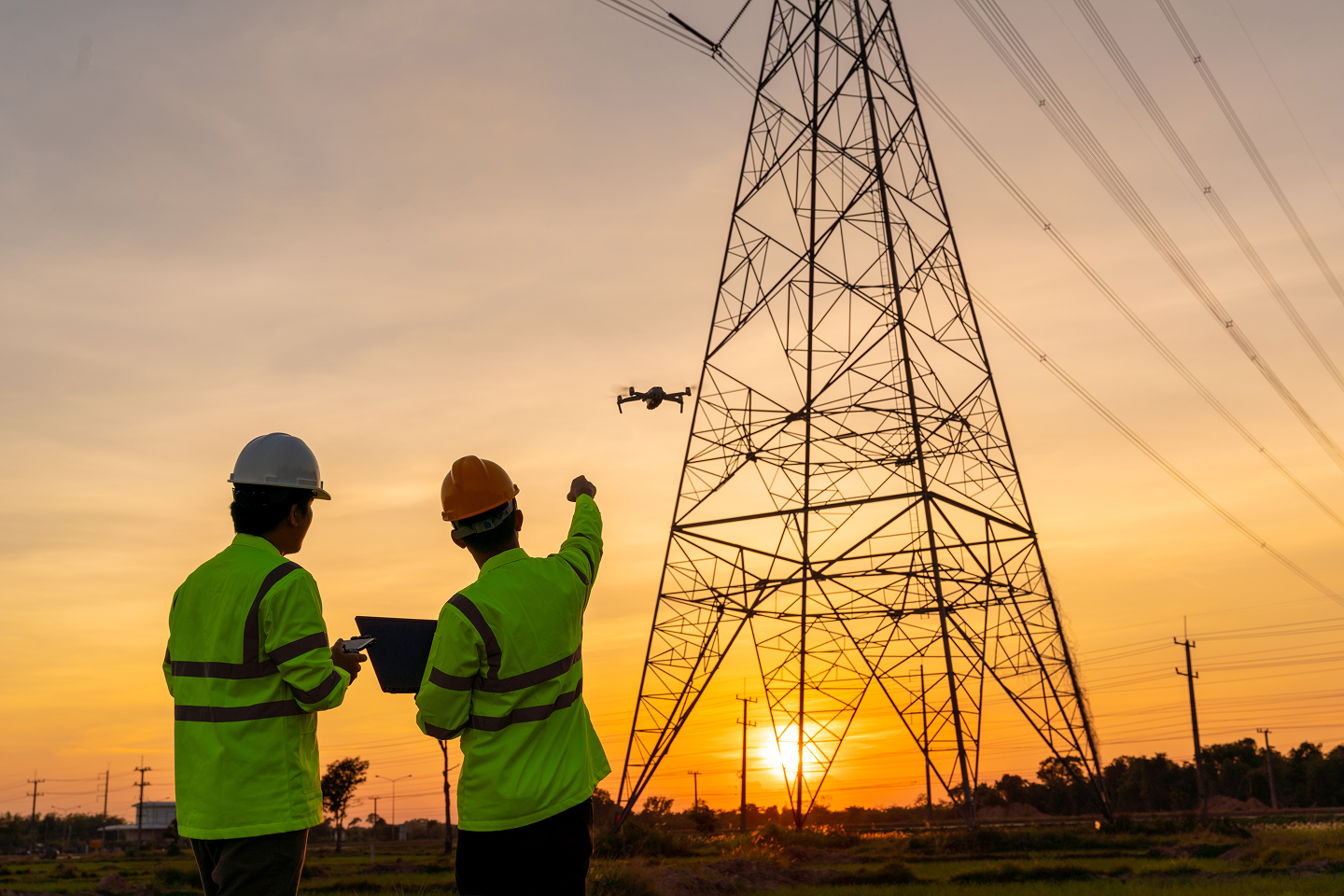 Two utility workers in hard hats flying a drone near a transmission tower at sunset