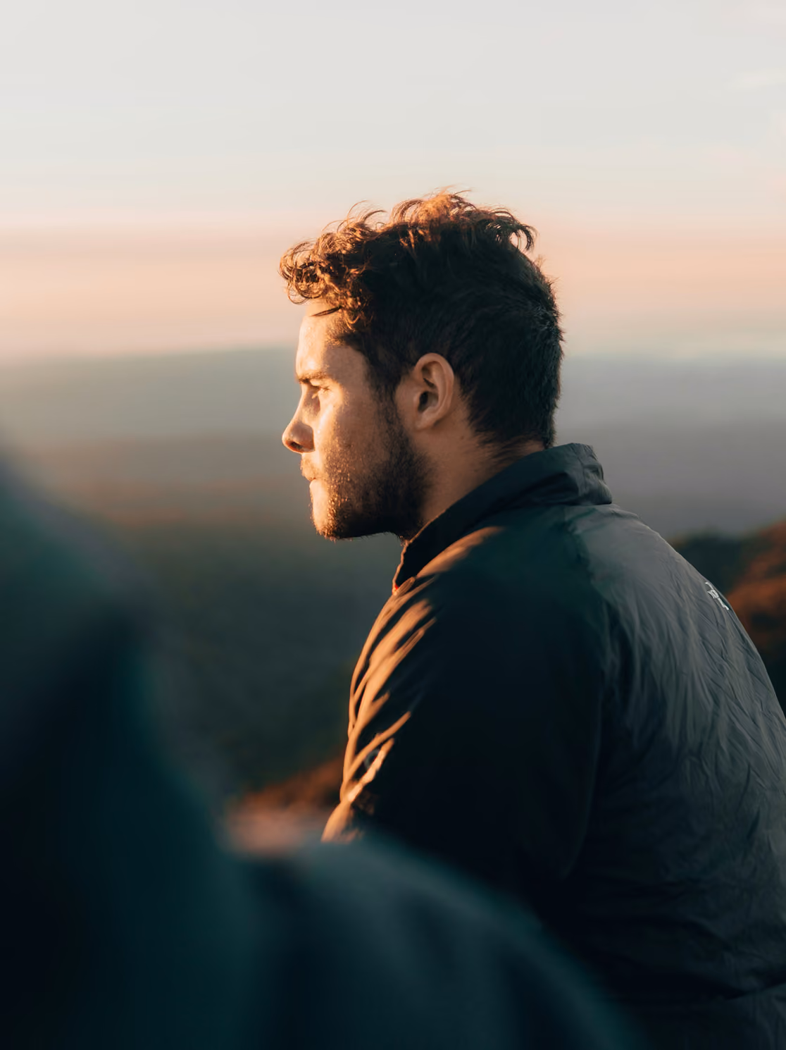 Side profile of a man with curly hair and beard looking contemplatively at the horizon during sunset, wearing a dark jacket with a blurred landscape in the background.