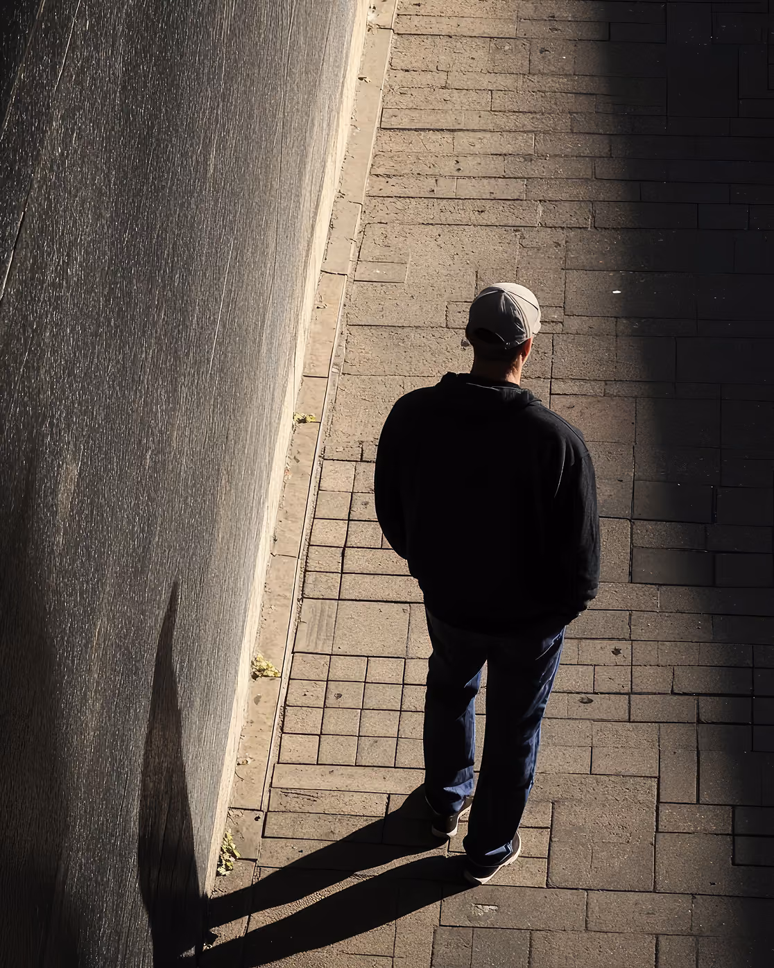 Person wearing a black jacket, blue jeans, and a light gray cap standing on a paved sidewalk near a wall with their back to the camera, casting a long shadow.