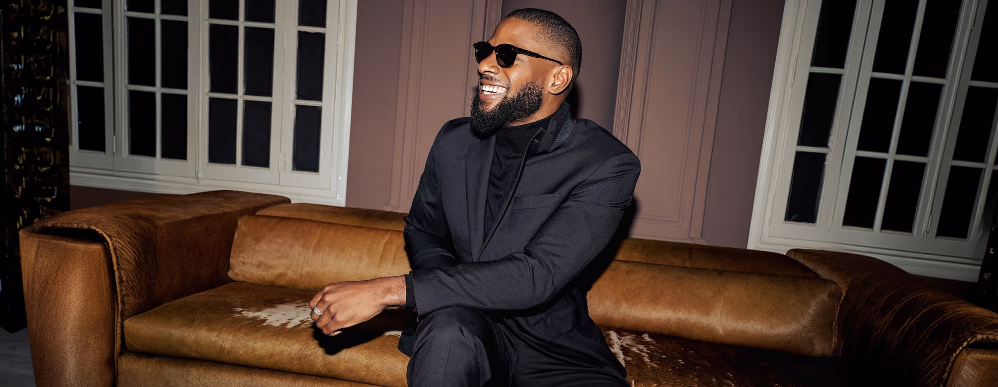Man in a black suit sitting on a leather couch, smiling
