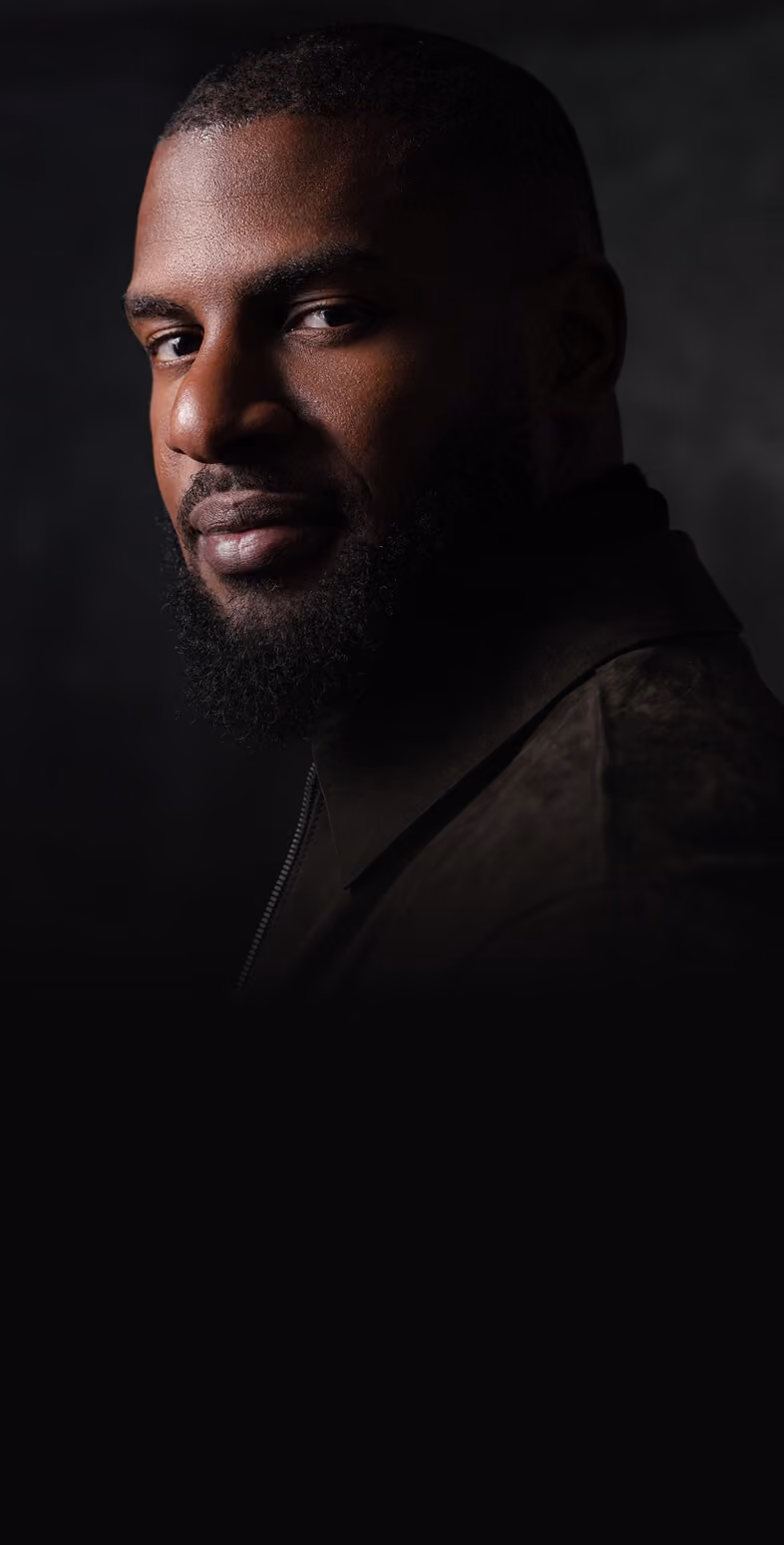 Profile portrait of a man with a short beard and close-cropped hair wearing black sunglasses and a black suit jacket against a dark background.