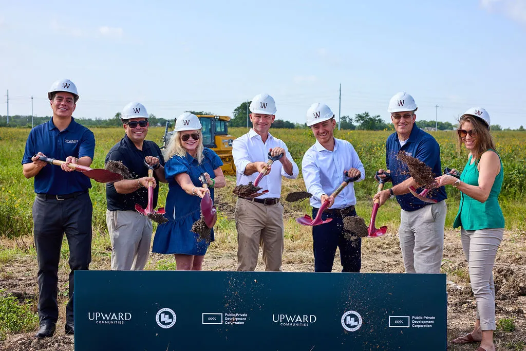 Upward Communities and public partner team members in hard hats standing on the raw Wildflower Lockhart site holding shovels of soil