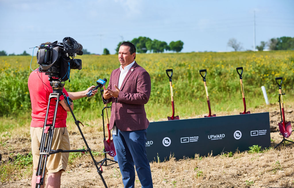 A speaker interviewed by a television news crew at the Wildflower Juniper Springs groundbreaking, with ceremonial shovels and partner banners behind him