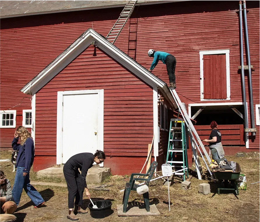 Neighbors repairing a red barn together — one on the roof, others mixing and hauling