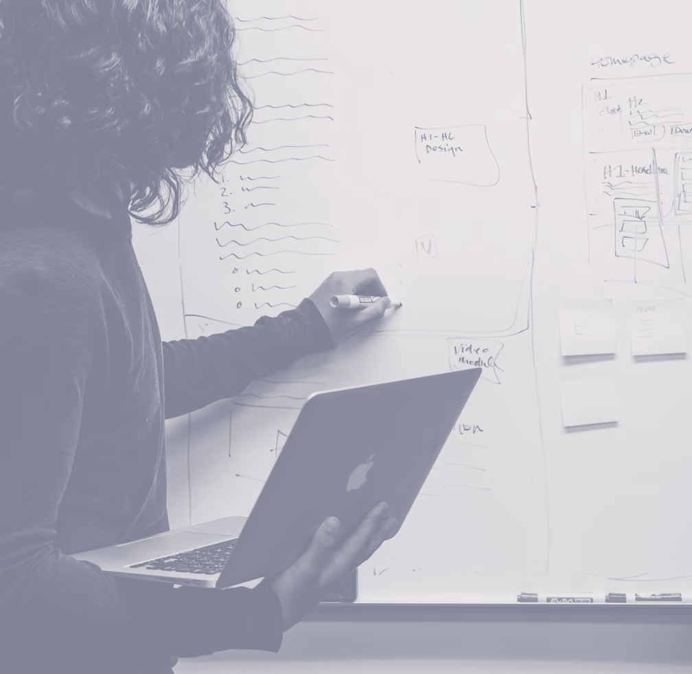 Person with curly hair holding a laptop and writing on a whiteboard covered in notes and diagrams with a marker.