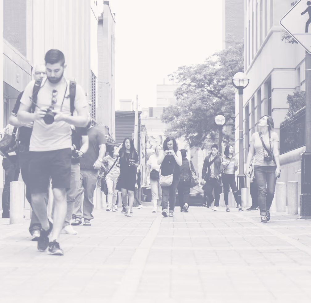 Pedestrians walking along a city street between buildings, some looking at cameras and phones while others walk forward.
