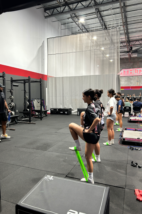 Young athletes participate in a sports performance class inside a large indoor training facility, with one athlete using a green resistance band while a coach instructs the group.