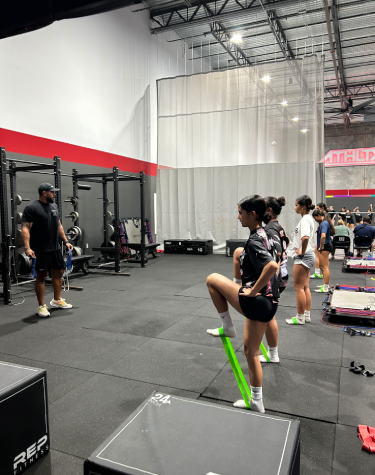 Young athletes participate in a sports performance class inside a large indoor training facility, with one athlete using a green resistance band while a coach instructs the group.