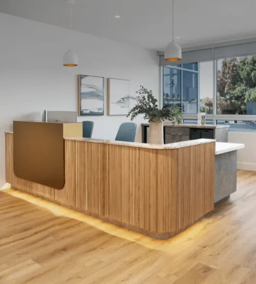 Modern reception area with a wooden front desk featuring under-lighting, green chairs behind the counter, pendant lights above, and large windows providing natural light.