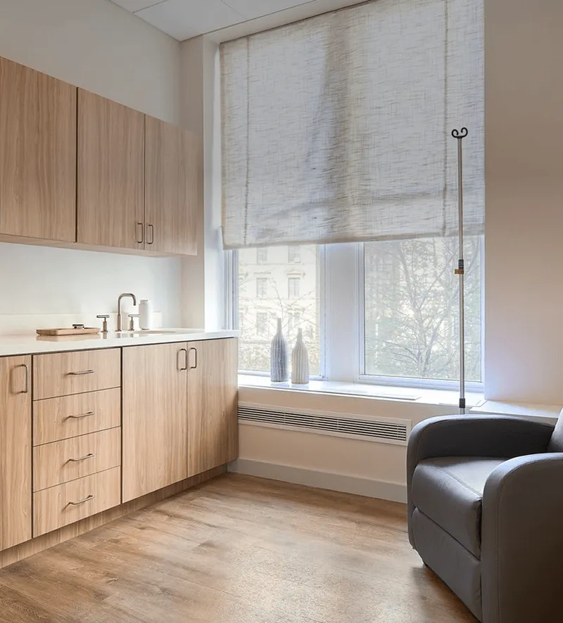 Bright medical consultation room with wood cabinets, countertop sink, and a comfortable gray recliner by a large window with sheer blinds.