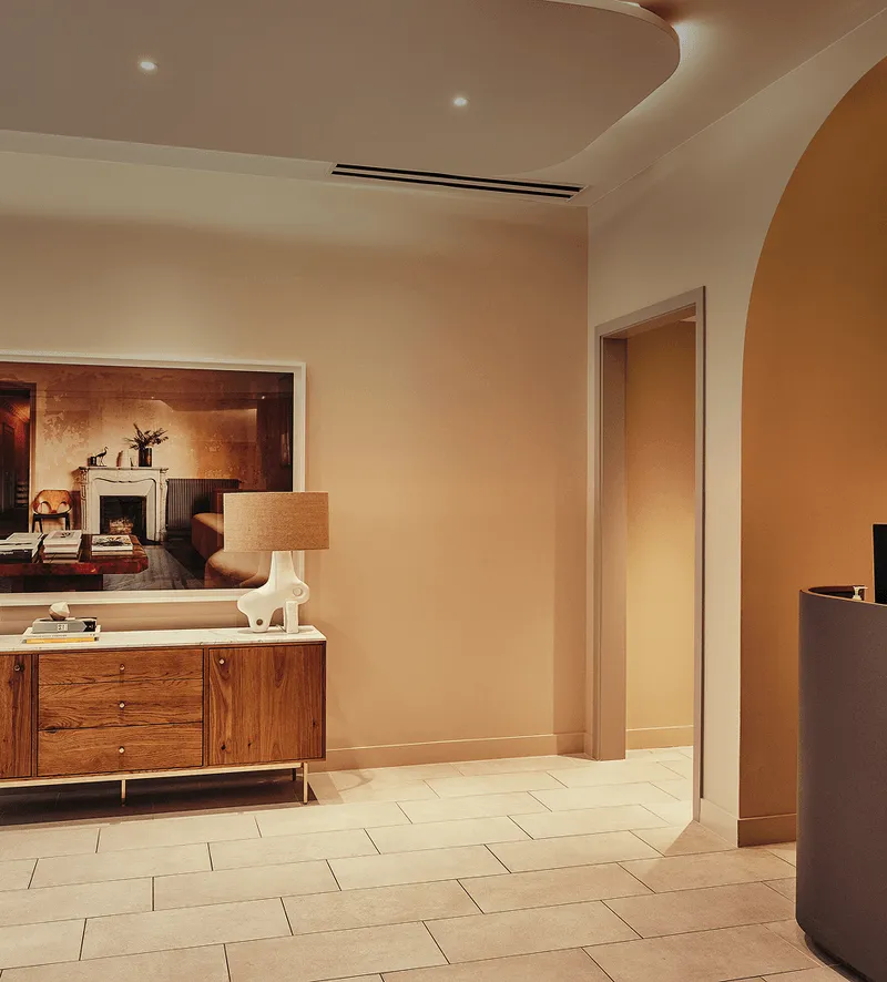 Modern interior space with warm beige walls, a wooden sideboard holding a lamp and books, and a large framed photo of a cozy room above it, featuring tiled flooring and an arched doorway.