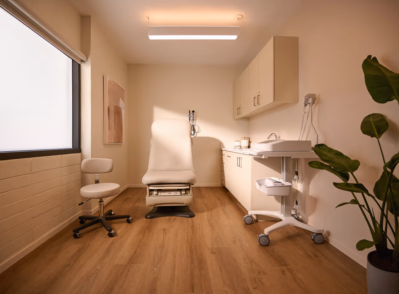Modern medical examination room with an empty exam chair, stool, sink, cabinets, and medical equipment, featuring warm lighting and a potted plant.
