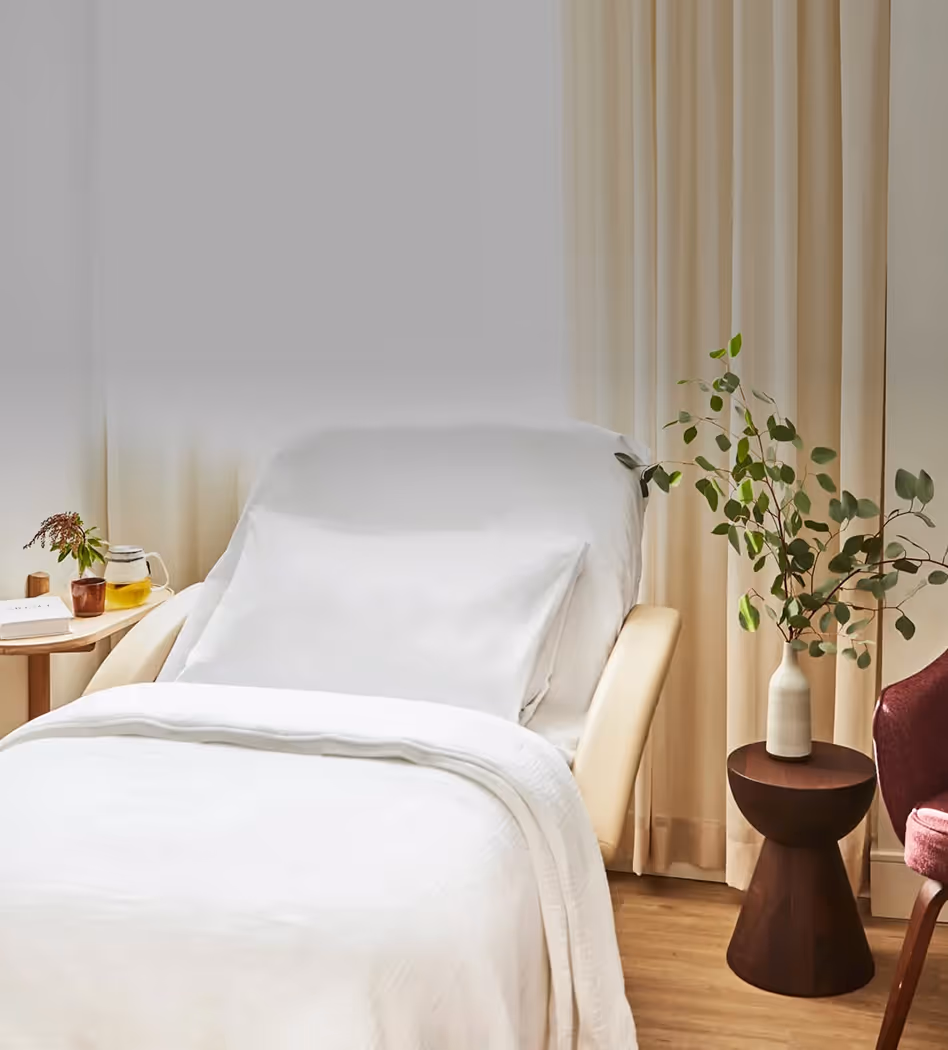 Bright medical or wellness room with a white treatment bed, built-in sink and cabinets, large window with natural light, and a view of trees and parked cars outside.