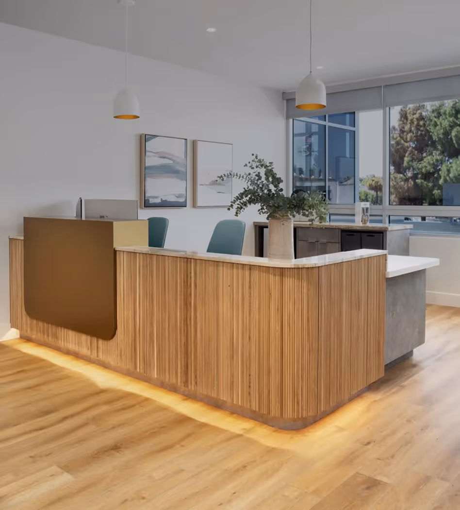 Modern reception area with a wooden front desk featuring under-lighting, green chairs behind the counter, pendant lights above, and large windows providing natural light.