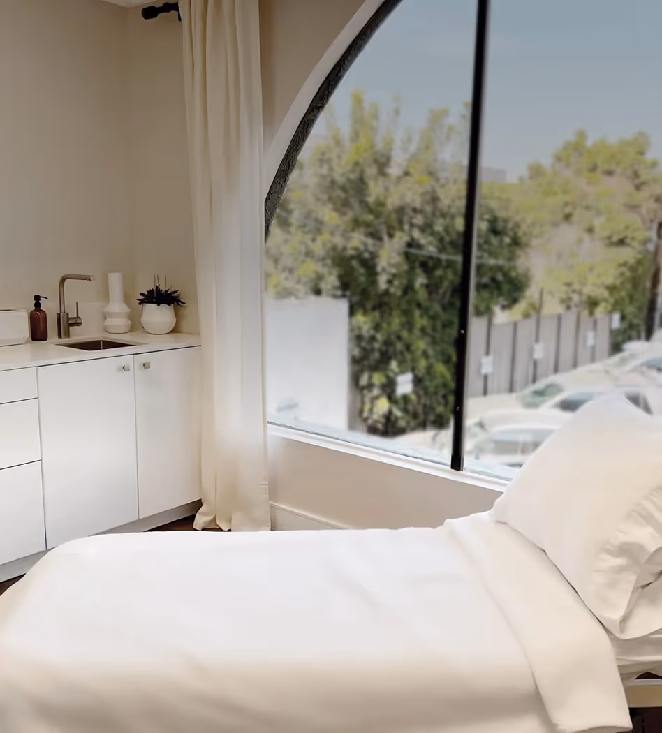 Bright medical or wellness room with a white treatment bed, built-in sink and cabinets, large window with natural light, and a view of trees and parked cars outside.