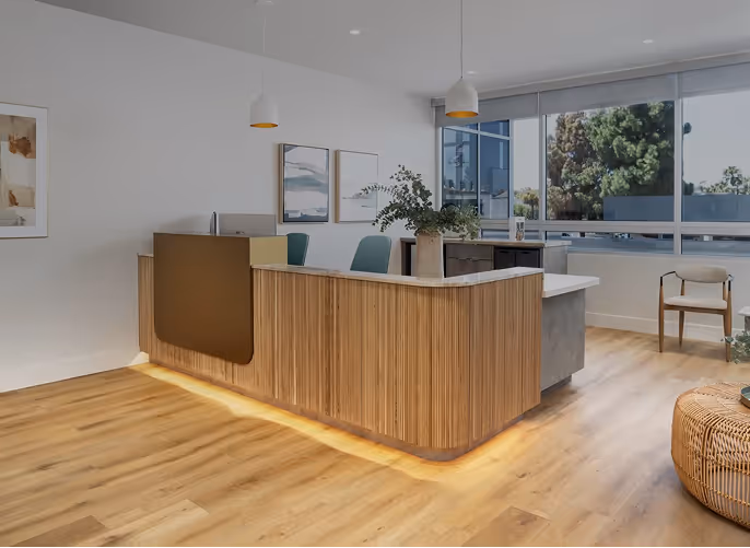 Modern reception area with a wooden front desk featuring under-lighting, green chairs behind the counter, pendant lights above, and large windows providing natural light.