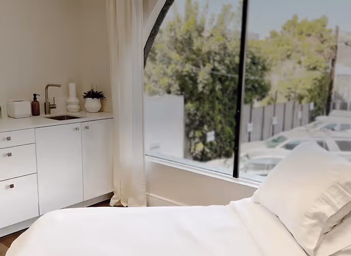Bright medical or wellness room with a white treatment bed, built-in sink and cabinets, large window with natural light, and a view of trees and parked cars outside.