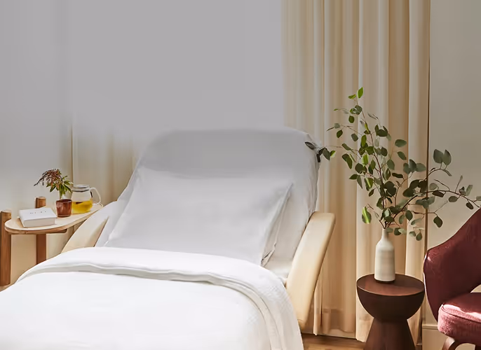 Bright medical or wellness room with a white treatment bed, built-in sink and cabinets, large window with natural light, and a view of trees and parked cars outside.
