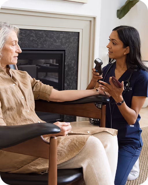 A healthcare professional uses a handheld medical device on an elderly woman's arm during a home visit.
