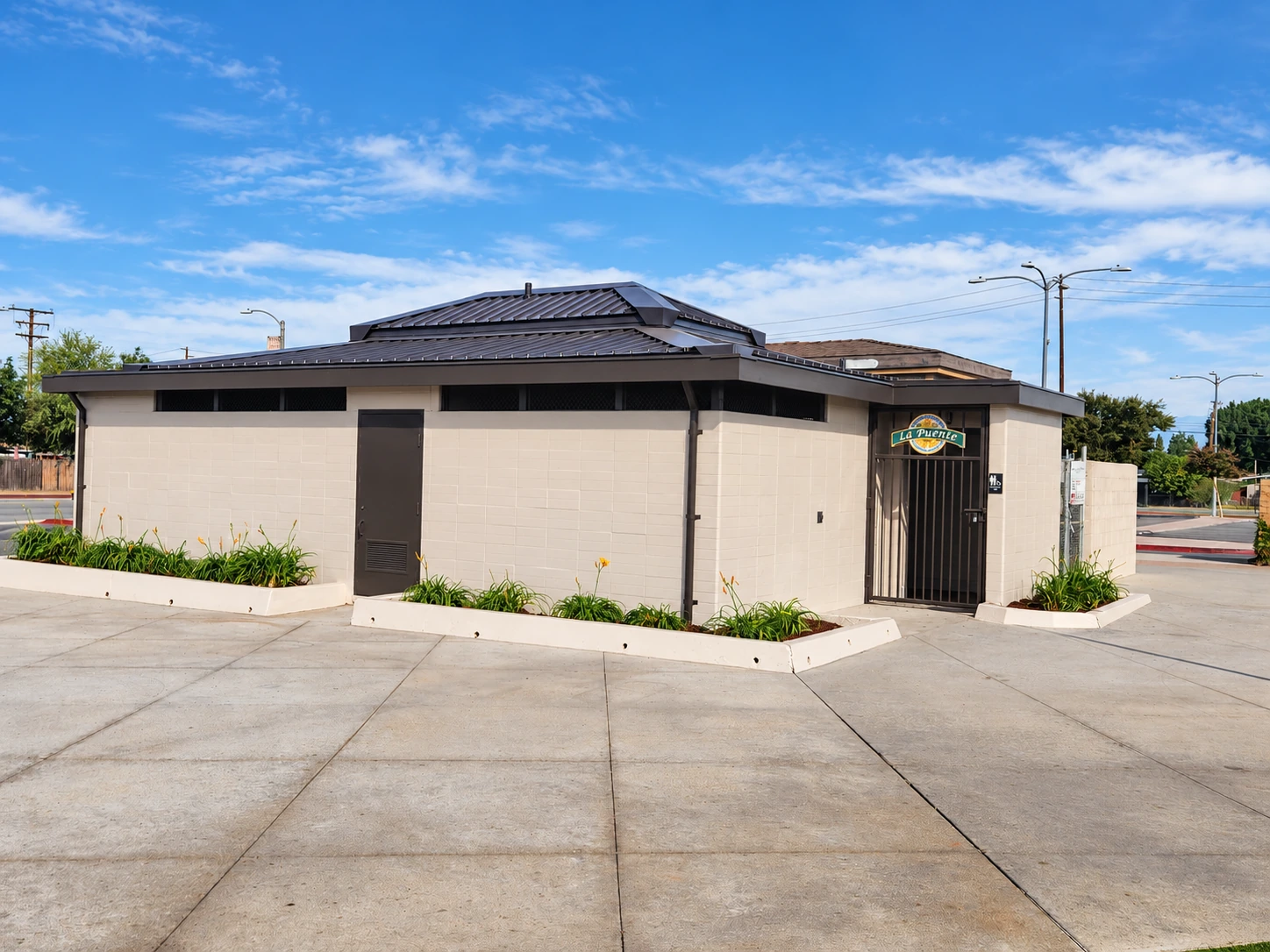 Completed Northwest Park restroom in La Puente with beige masonry walls, a dark roof, and a gated entrance.