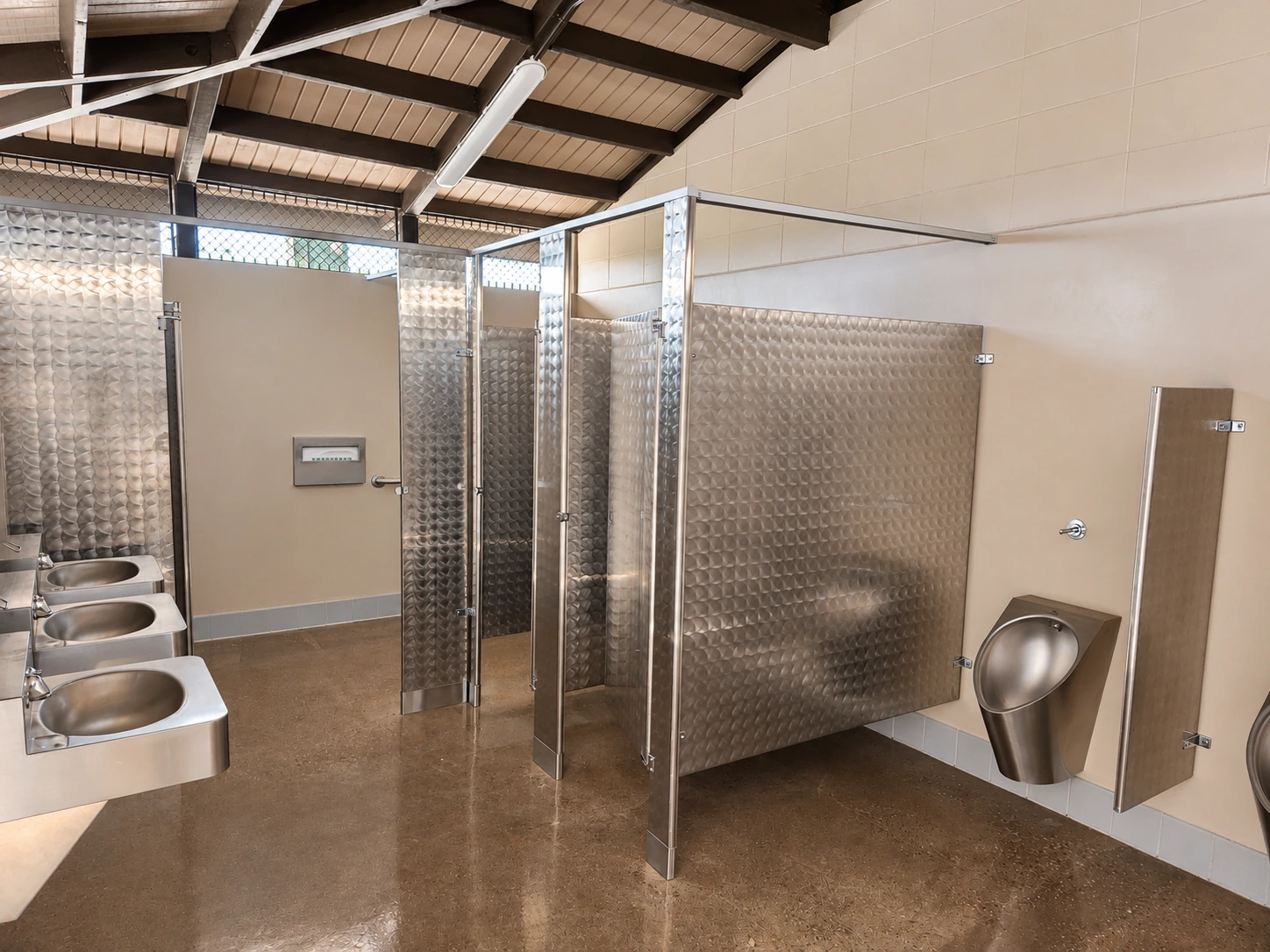 Renovated Northwest Park restroom interior with stainless steel partitions, plumbing fixtures, sealed concrete flooring, and exposed roof framing.