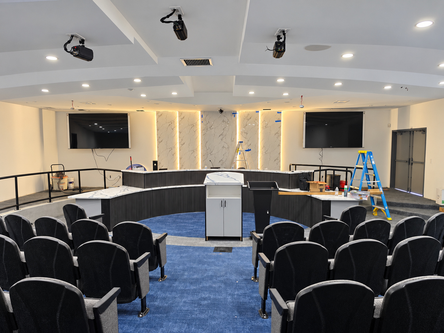 Front view of the Adelanto Council Chamber nearing completion, showing the curved dais, public seating, illuminated wall panels and remaining construction tools.
