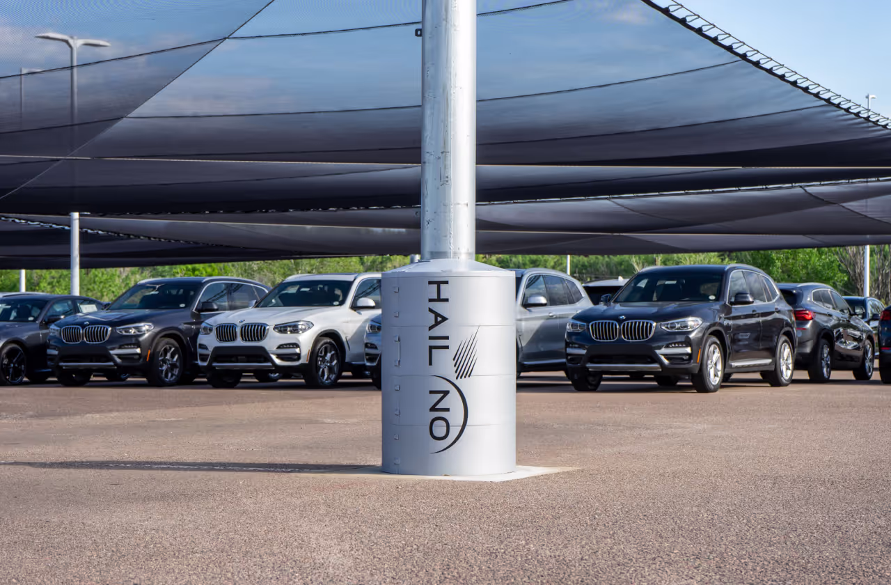 BMW SUVs parked under a Hail No netting canopy at a dealership, with a branded support column in the foreground.