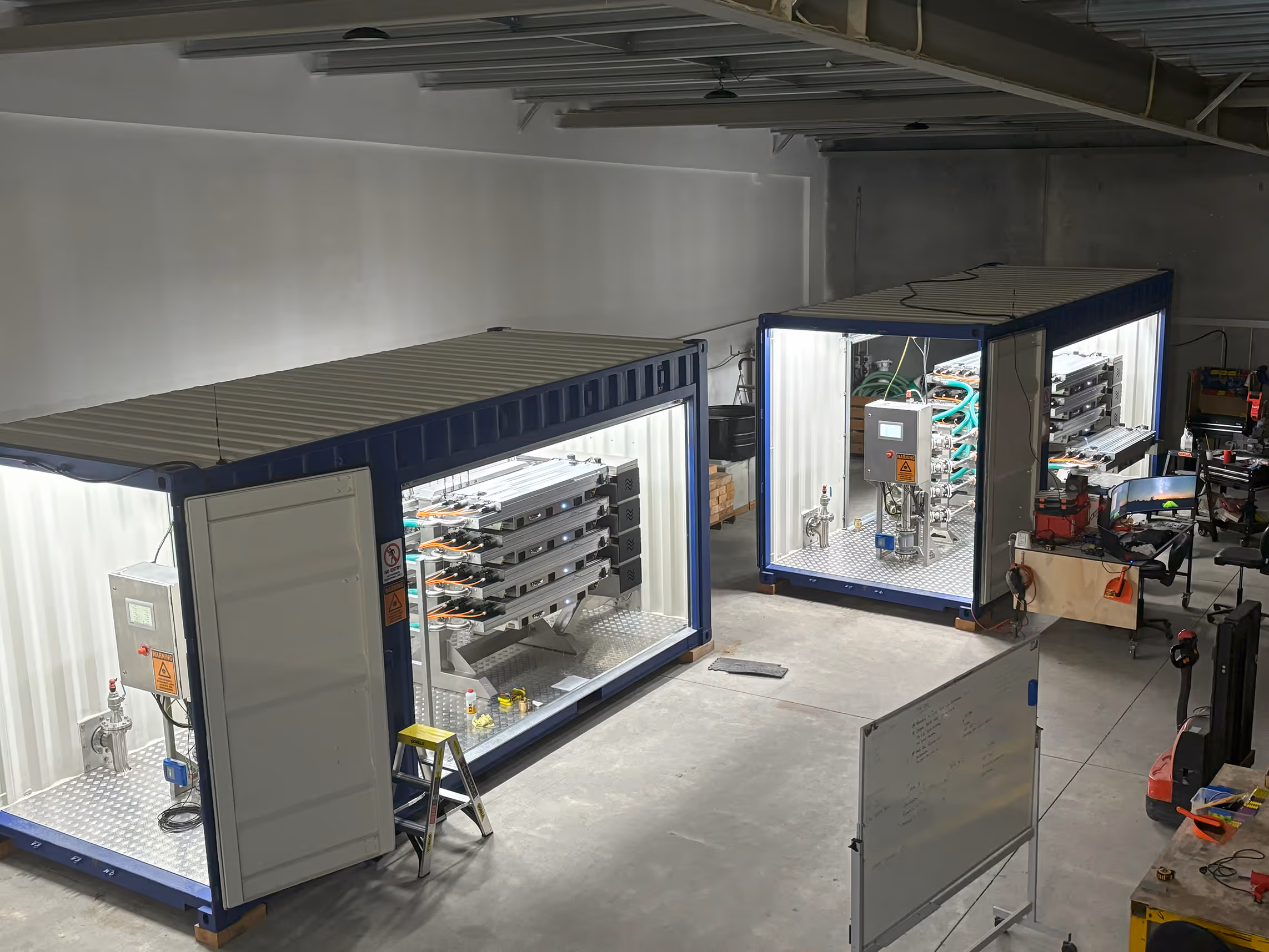 Two blue shipping containers with industrial piping and control panels inside a warehouse.