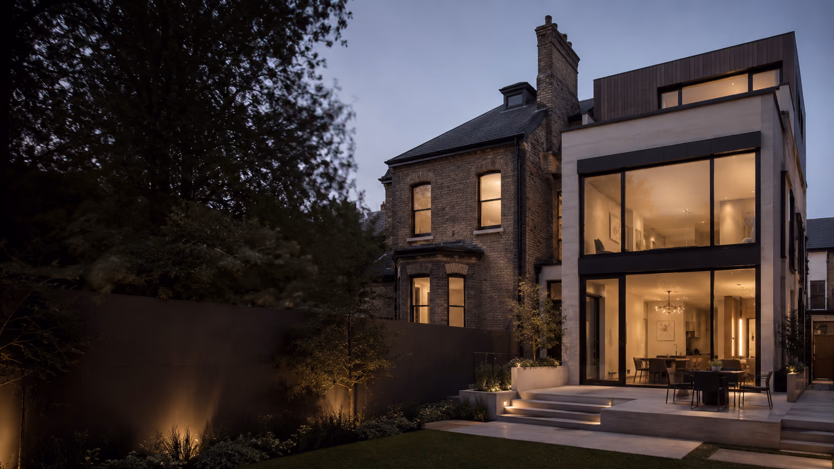 Modern house extension with large glass windows illuminated at dusk, next to an older brick building.