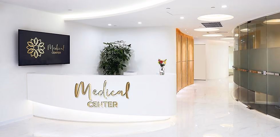 Bright medical center reception with white desk, gold lettering, plant, flowers, and curved glass walls.