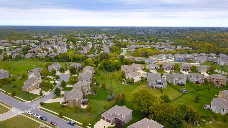 Aerial view of a suburban neighborhood with houses, green lawns, trees, and roads under a cloudy sky.