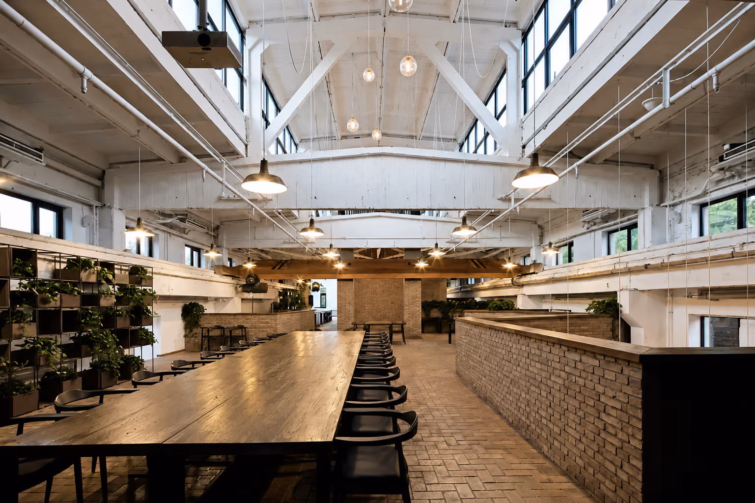 Spacious industrial-style interior with a long wooden table surrounded by black chairs, exposed white beams, hanging lights, and large windows allowing natural light, complemented by brick walls and plants on the left side.