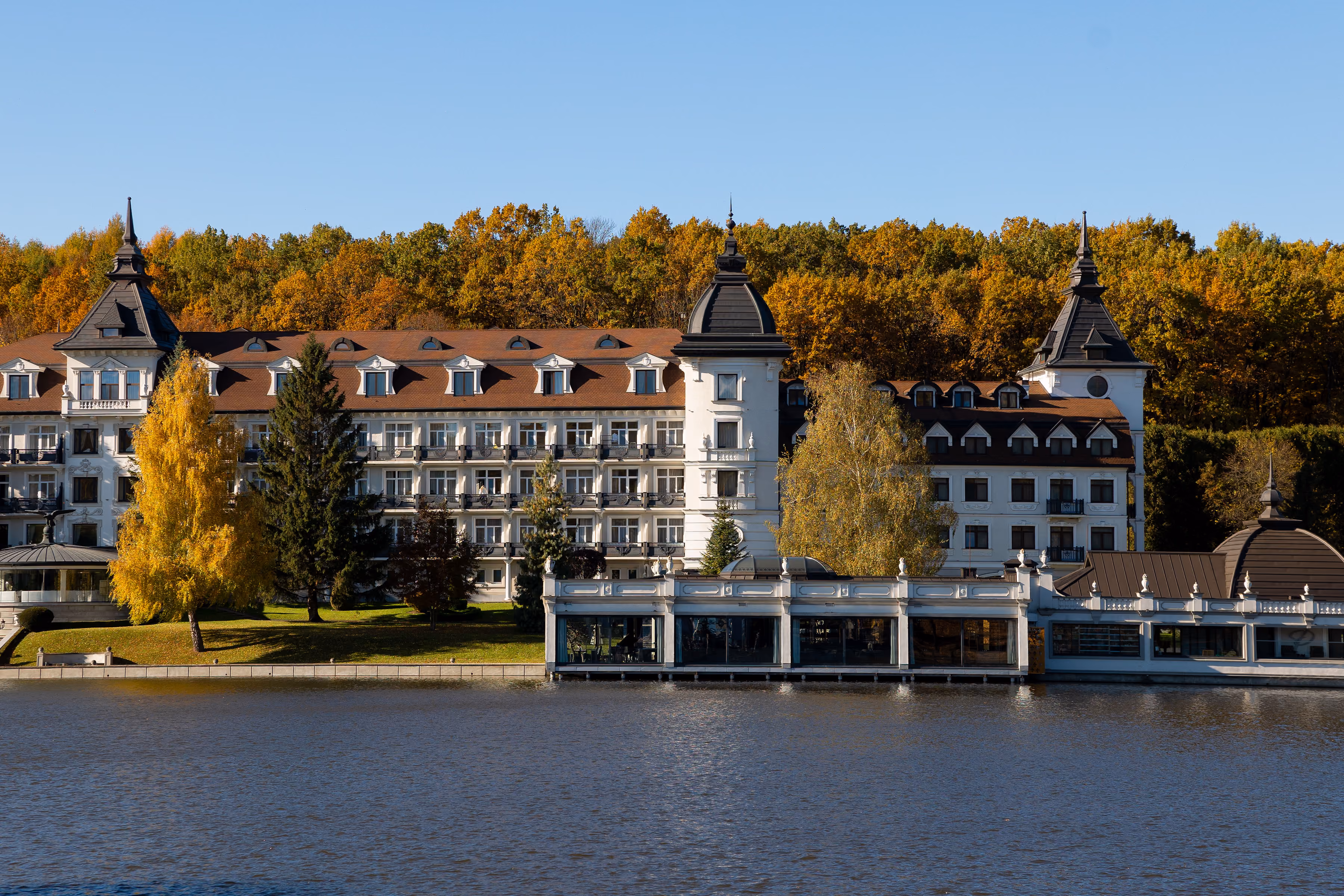 Classic large building with towers by a lake and autumn trees in the background under clear sky.