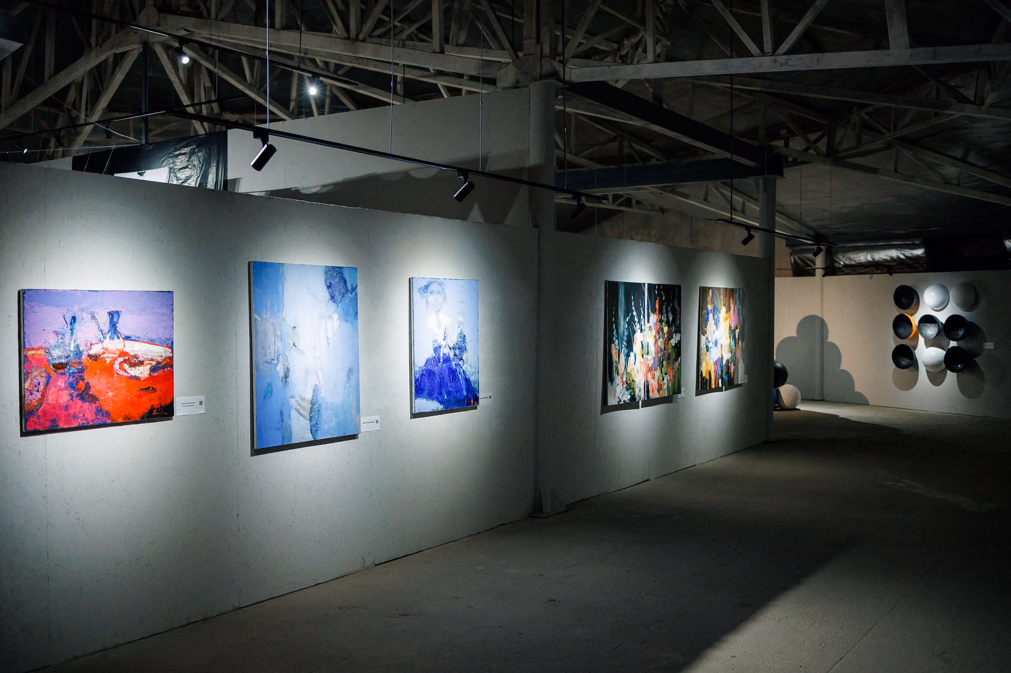 Gallery wall shows six abstract paintings lit by spotlights in an industrial-style space.