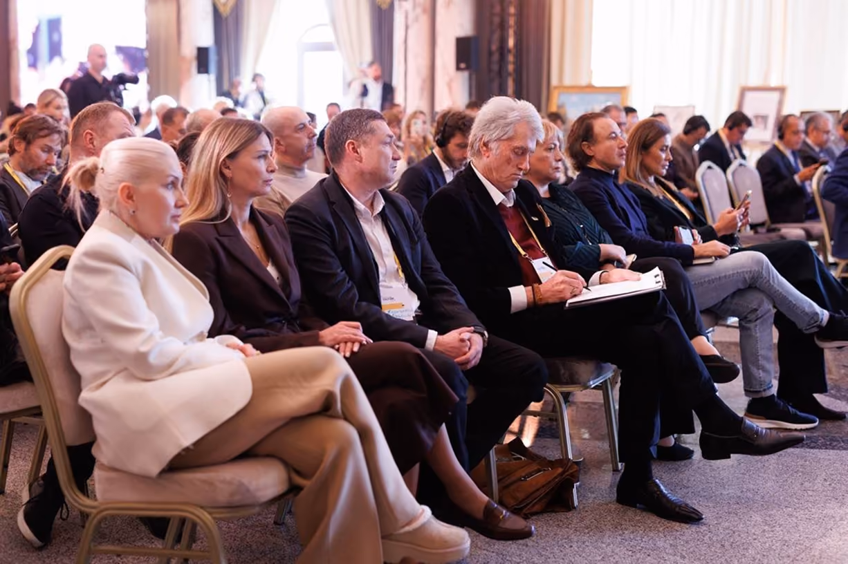Audience of men and women sitting in rows at a conference, attentively listening and taking notes.
