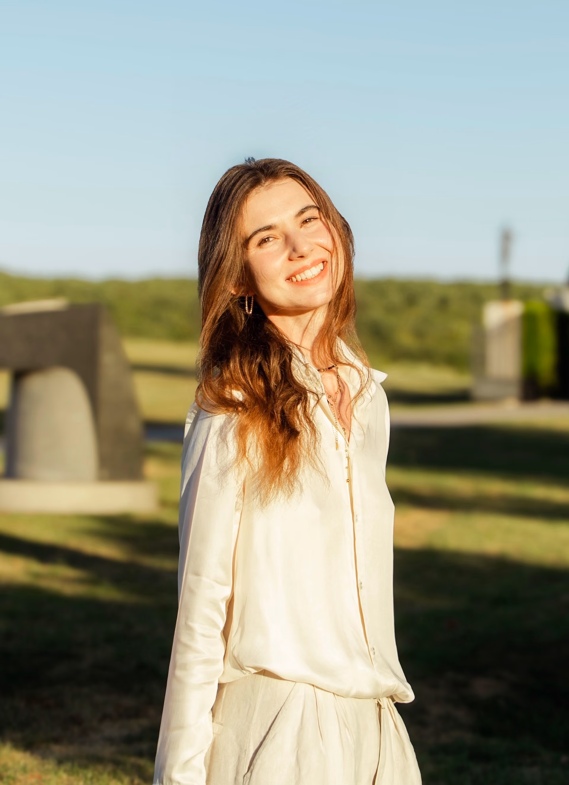 Young woman with long hair smiling outdoors in sunlight wearing a light-colored blouse and pants.