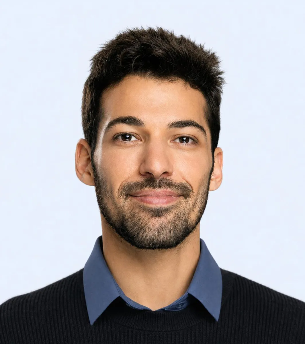 Young man with short dark hair and beard wearing a blue-collared shirt under a black sweater.