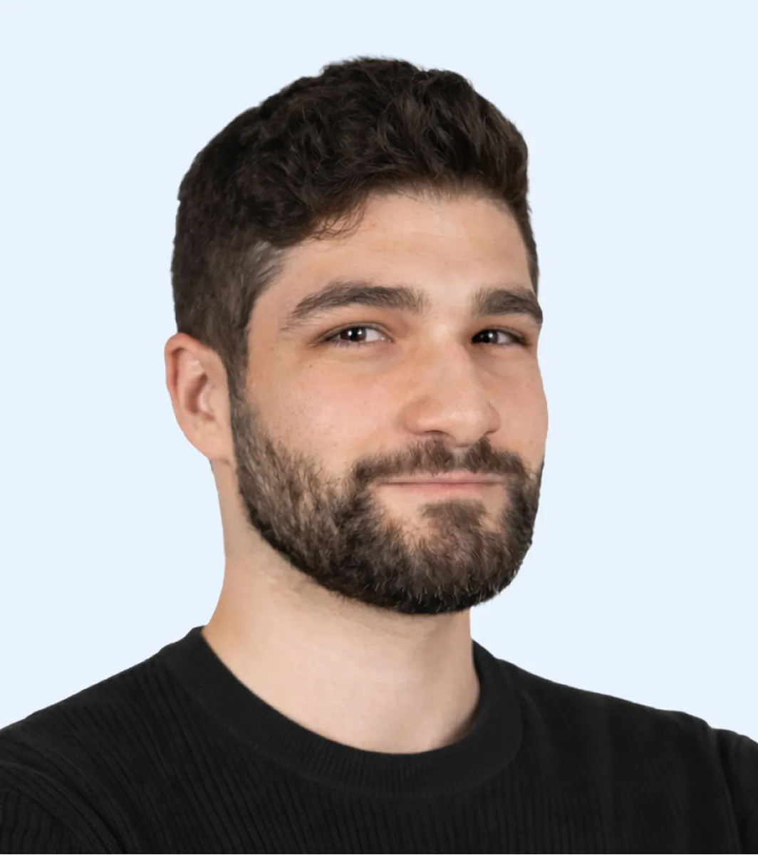 Man with dark hair, beard, and black sweater smiling against light blue background.