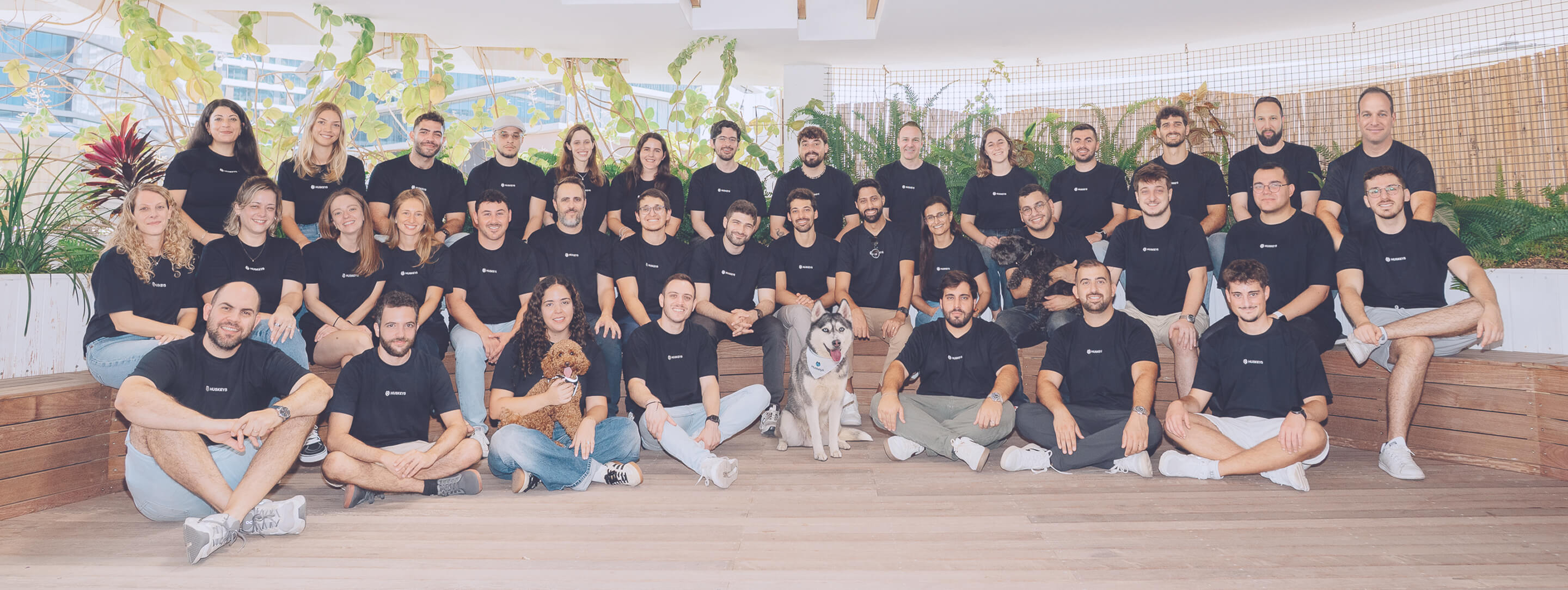 Group of 30 people in black t-shirts with logos seated on wooden benches surrounded by plants indoors.