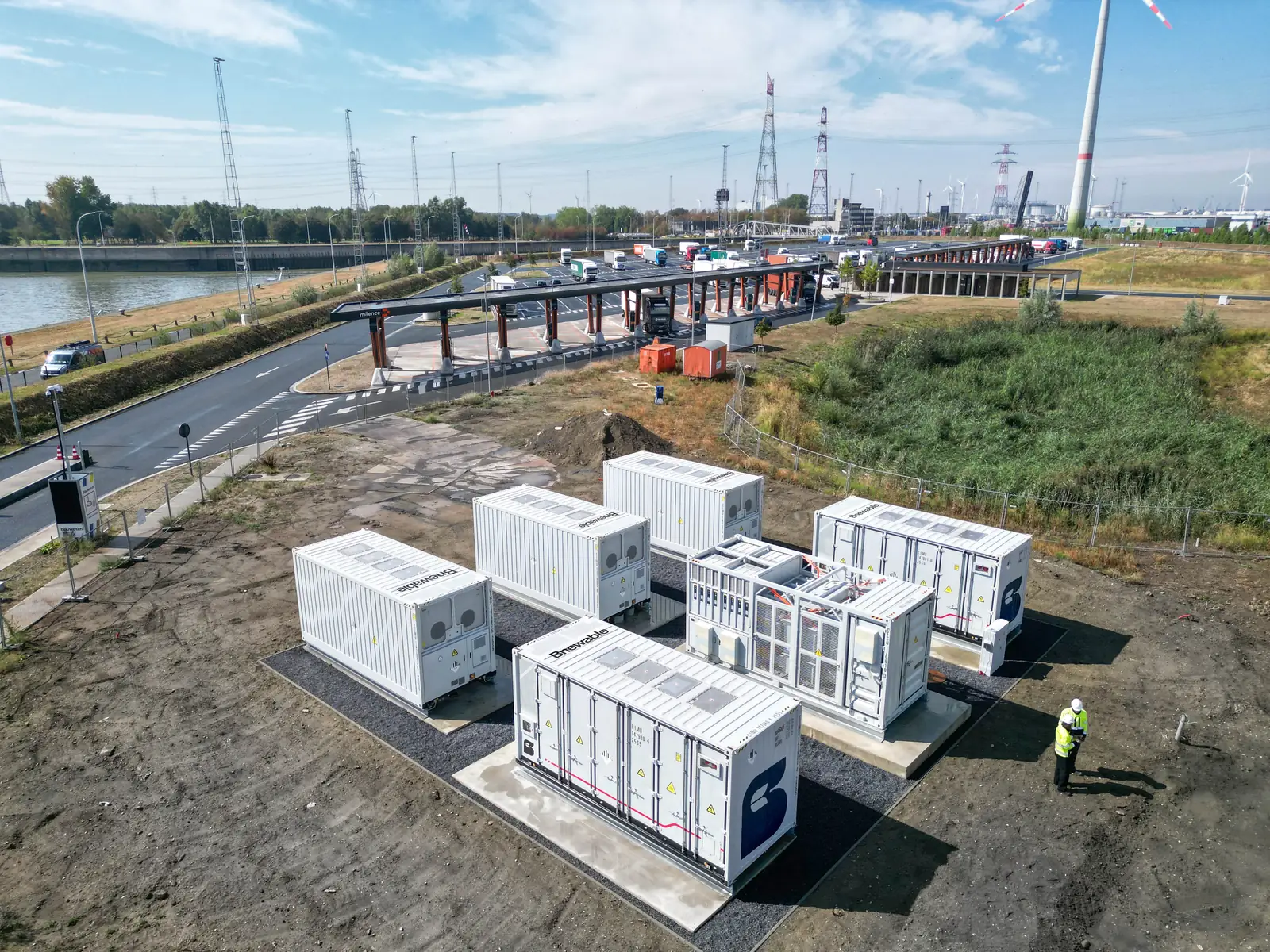 Aerial view of five Bnewable battery storage containers and an inverter skid installed beside an electric truck charging hub, with a technician on site and wind turbines on the horizon